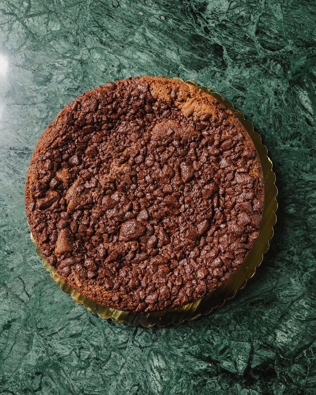Chocolate Chip Cookie Cake entero, cenital sobre su tarima dorada en el mármol verde del café